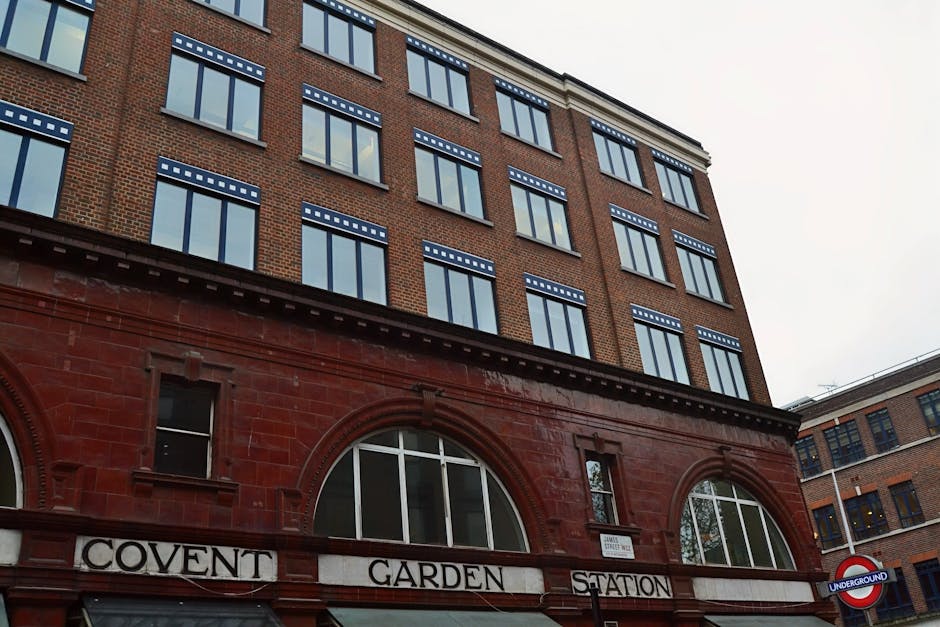 A busy urban street scene in Covent Garden with a row of multi-story buildings featuring detailed architectural facades, large windows, and decorative elements, illuminated by warm natural sunlight. T