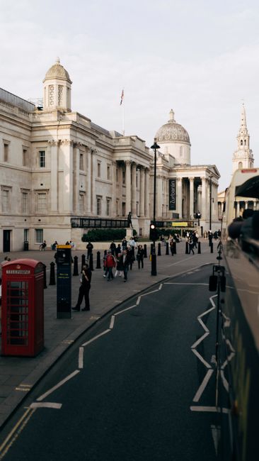 A wide view of Trafalgar Square in Westminster, London, showing a large granite statue of a historical figure with a draped cloak, standing on a tall rectangular pedestal in the foreground. To the lef